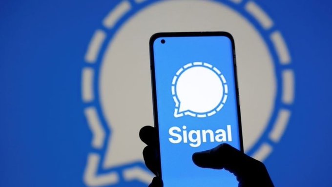 A hand holds a smartphone displaying the Signal app logo against a blurred blue background featuring the same logo.