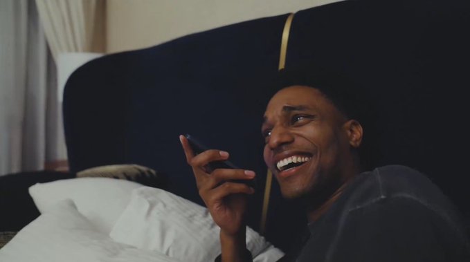 A young man lying on a bed smiles and laughs while holding a phone to his ear.