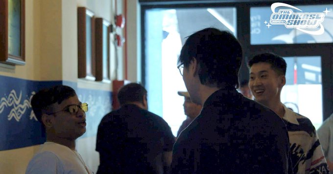 A group of men chat in a hallway, with one wearing yellow-tinted glasses in the foreground, while a watermark in the top right reads "THE OMAKASE SHOW." The image appears to capture the casual gathering at The Omakase Show during Token2049 in September 2024, where the artist @kolahonn was met.