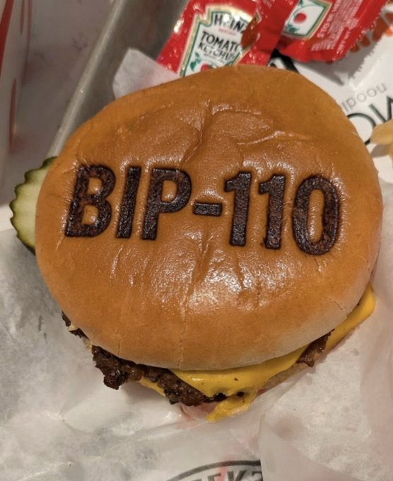 A close-up of a hamburger bun stamped with 'BIP-110' sitting on a tray with ketchup packets and pickles.