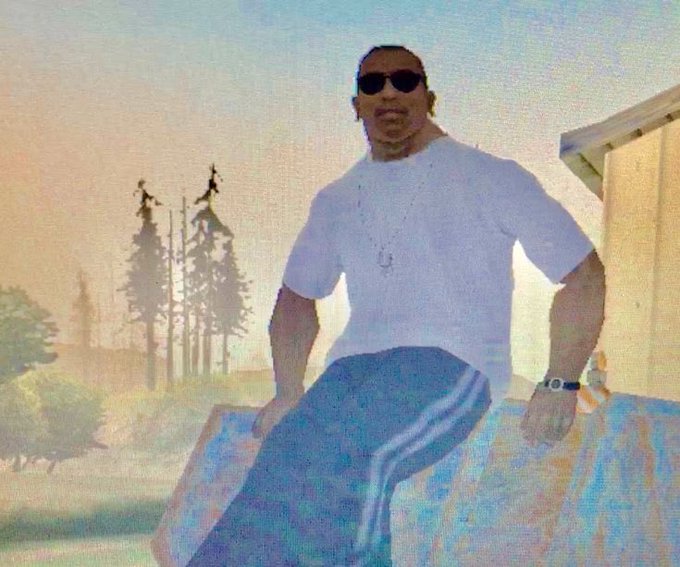 A grainy, stylized image shows a man wearing sunglasses, a white t-shirt, and blue track pants with white stripes, sitting outdoors with a hazy, dreamlike background featuring trees and a building.