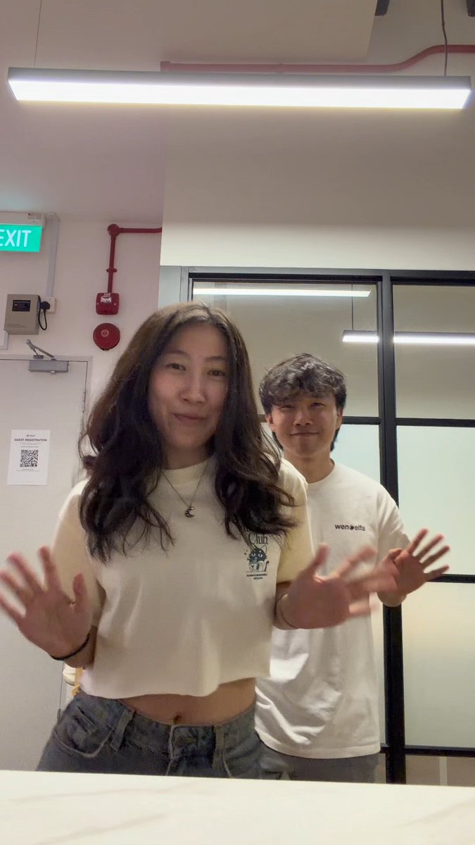 Two casually dressed young people pose indoors under fluorescent lighting, with the woman in front wearing a white cropped t-shirt and the man behind in a white shirt reading "wen belts," both smiling and gesturing towards the camera; a green "EXIT" sign and a "GUEST REGISTRATION" QR code are visible on the wall.