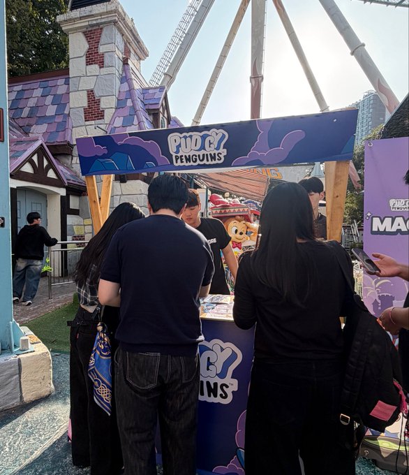 People gathered at an outdoor 'Pudgy Penguins' promotional booth near what appears to be a roller coaster or theme park ride.