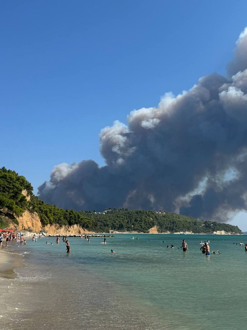 A crowded beach with swimmers enjoying turquoise waters, while a massive plume of dark wildfire smoke billows ominously over the nearby hills in the distance. The image captures the juxtaposition of a relaxing seaside vacation in Lefkada, Greece, with the unsettling reality of having to escape a wildfire, mirroring the post's tone of finding unexpected profits amid chaotic circumstances.