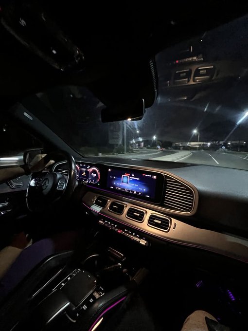 An interior view from the passenger seat of a Mercedes-Benz vehicle at night shows the illuminated dashboard with digital displays, air vents, and ambient lighting, with the road and city lights visible through the windshield.