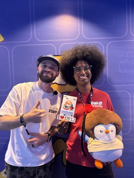 Two people pose together holding merchandise; one wears a white shirt and cap giving a thumbs-up, while the other wears a red 'Studio Job' shirt and holds a plush penguin toy, against a purple backdrop with a 'BYL' card visible.