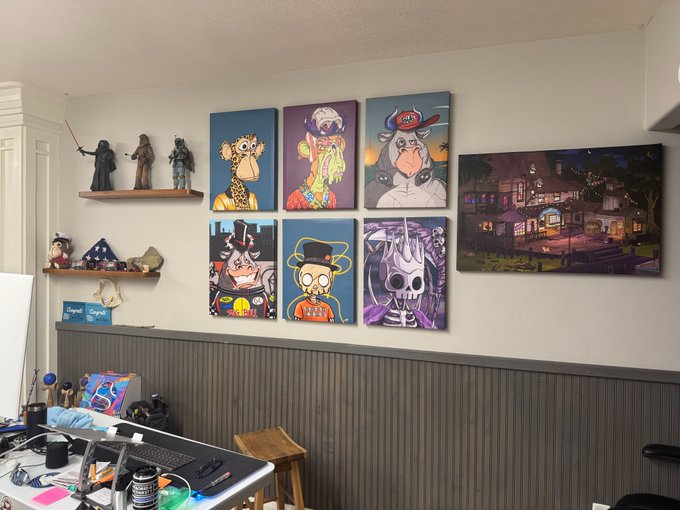 A home office or studio space features walls decorated with framed cartoon art prints (including giraffes, skulls, and characters), Star Wars figures on floating shelves, a desk with computer equipment, and a separate framed artwork of a colorful building.