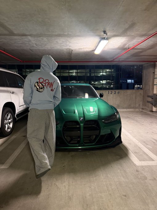 A person wearing a gray hoodie with a "Brain" logo stands in a parking garage facing the front of a green BMW M4 next to a white SUV.