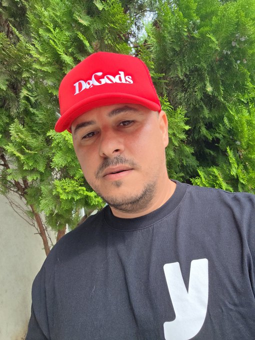 A man wearing a red 'DeGods' cap and a black t-shirt poses for a selfie in front of green coniferous foliage.