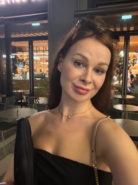 The image features a woman wearing a black dress standing outside of a restaurant or cafe at night. She has her arm around herself as she poses for a picture with a smile on her face. The background includes several chairs, dining tables, and potted plants, suggesting that this is an outdoor seating area. There are also other people present in the scene, but they are not the main focus of the image.