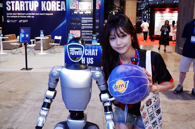 A young woman posing with a silver humanoid robot at the 'Startup Korea' exhibition, holding a blue basketball with 'SEJONG' text, with event signage visible in the background.