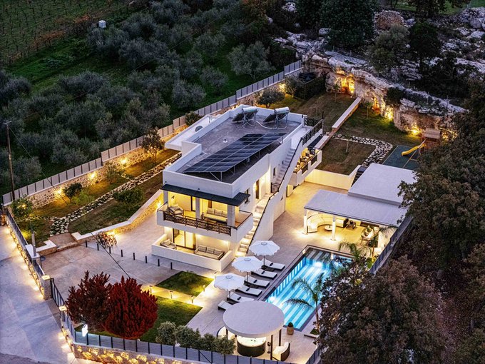 An aerial view of a luxurious modern villa at night, featuring a swimming pool, sun loungers, and solar panels on the roof, surrounded by landscaped gardens.