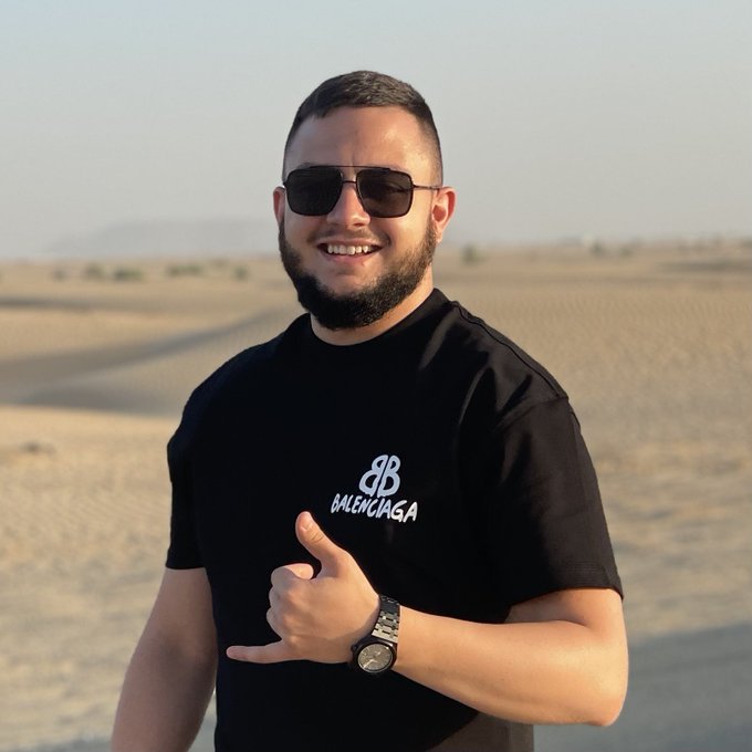 A man with a beard and sunglasses wearing a black Balenciaga t-shirt, giving a thumbs-up gesture while standing in a desert landscape.