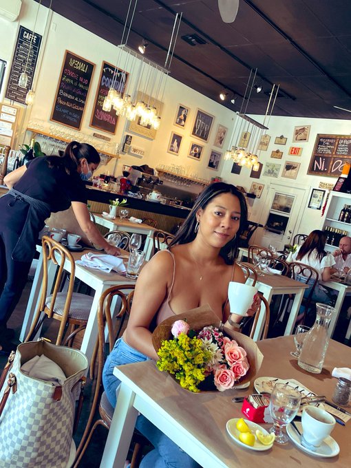 A woman with long dark hair sitting at a table in a cafe, holding a bouquet of yellow, pink, and purple flowers and a white coffee cup, with a checkered Louis Vuitton-style bag beside her.