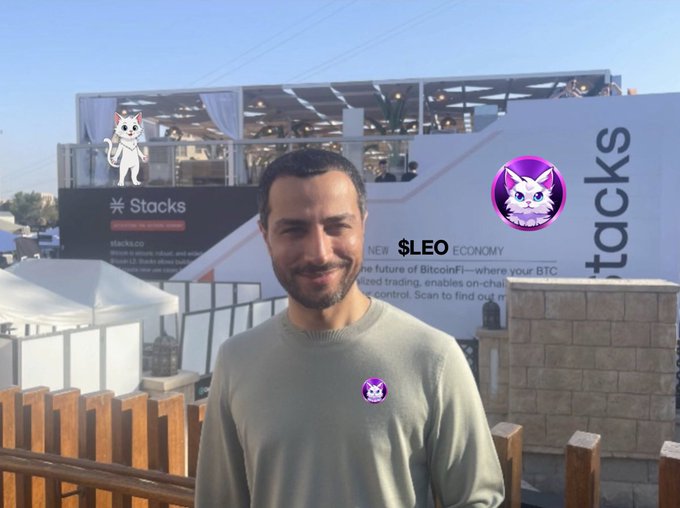 A man in a light green sweater standing outdoors in front of a 'Stacks' booth promoting 'Bitcoin L2 trading' and a 'NEW $LEO ECONOMY'.