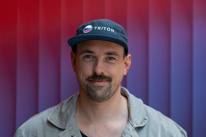 Portrait of a smiling man wearing a gray 'Triton' Pepsi-branded cap and olive shirt against a red-to-purple gradient background.