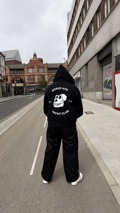 Person wearing a black 'BORED APE YACHT CLUB' hoodie standing on a city street with buildings in the background.