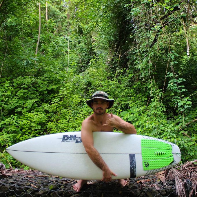 A shirtless man in a camouflage bucket hat holds a white surfboard with a green traction pad, standing barefoot in front of lush jungle foliage. The caption promotes forming a "Solana surf team," tying the surfer's adventurous lifestyle to promoting the Solana crypto community and its members.