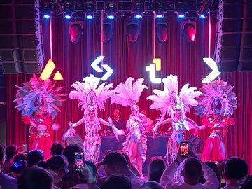 The image features a group of people dressed up as dancers performing on stage under bright lights. The performers are wearing feather boas and other costumes that make them look like they're dancing at a club or concert. There are several people standing around, watching the performance with interest. In addition to the dancers, there is also a cell phone visible in the scene, possibly being used by someone in the audience to capture the moment or share it on social media.