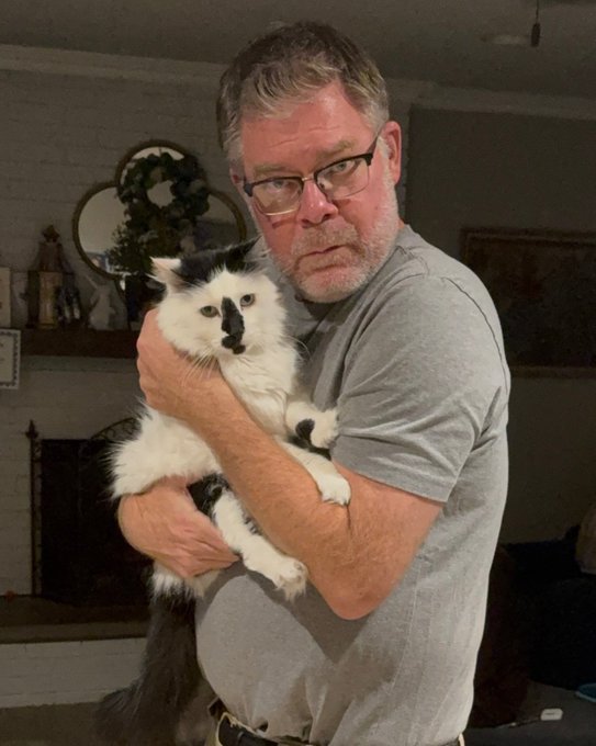 A man with glasses and a gray t-shirt holds a fluffy black and white cat in what appears to be a home interior.
