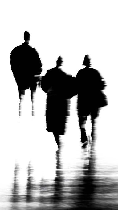 A blurry, black-and-white photograph showing the silhouettes of three people walking together, their forms and reflections distorted by motion blur.