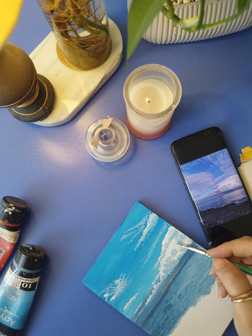A person's hand uses a small paintbrush to paint a blue seascape on a small canvas, with a lit candle, paint tubes, and a smartphone displaying a reference image of the sea nearby on a blue surface.