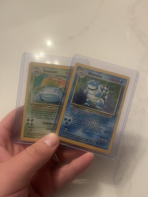 A hand holding two Pokemon trading cards in protective sleeves: a Venusaur and a Blastoise, both first edition holographic cards.