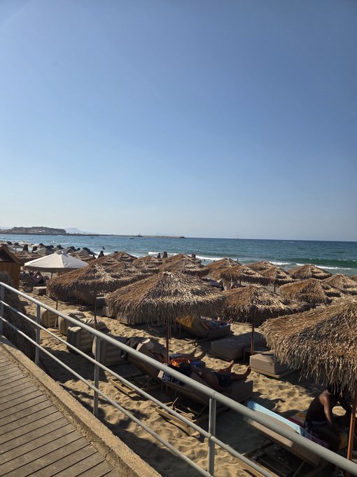 A sunny beach scene viewed from a wooden boardwalk, showing rows of straw umbrellas shading lounge chairs along the sandy shore with the blue ocean and a small island in the distance.
