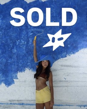 A woman in a yellow outfit holds a paintbrush and a mask-shaped palette in front of a wall painted blue and white with the word 'SOLD' displayed prominently.