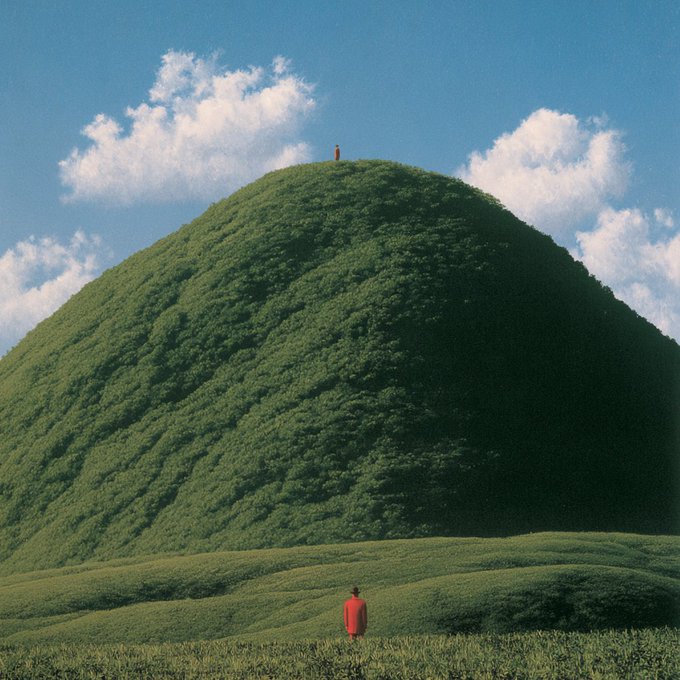 A surreal landscape showing a small person in a red coat standing in a grassy field looking at a massive green dome-shaped hill, with another small figure standing at the peak of the hill under a blue sky with scattered clouds.