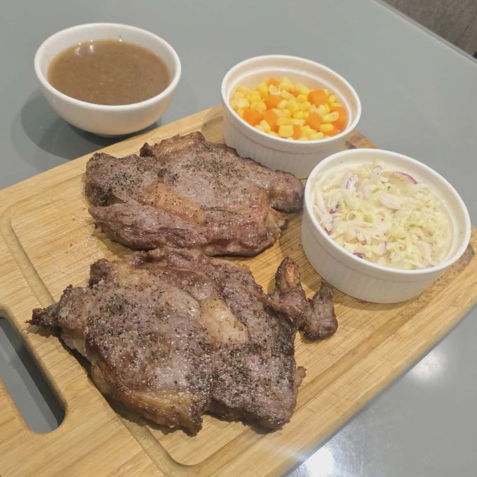 A meal served on a wooden cutting board featuring two seasoned steaks, a bowl of gravy, a ramekin of diced carrots and corn, and a ramekin of creamy coleslaw.