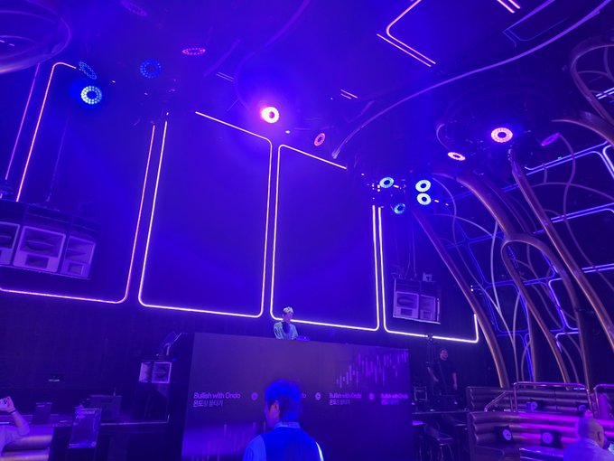A dimly lit nightclub or concert venue is illuminated by blue and purple neon lights with a DJ performing on an elevated stage while patrons watch below.