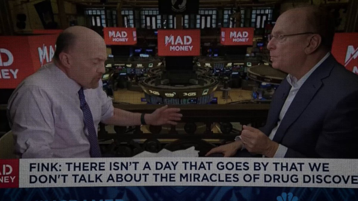 The image shows two men having a conversation, with one man wearing glasses and the other wearing a suit. The background features a news broadcast setting with multiple TVs displaying various news stories. One of the TVs has the text "Fink: There isn't a day that goes by that goes by that we don't talk about the miracles of drug discovery." This quote is from a post on Twitter, which is related to the image and the conversation between the two men.