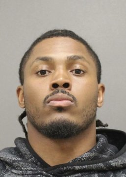 Mugshot-style photograph of a young man with braided hair and a beard wearing a gray patterned hoodie against a plain gray background.