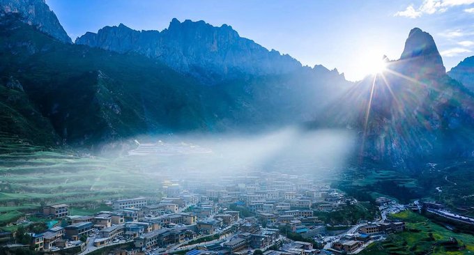 An aerial view of a mountain valley village shrouded in mist, with sunlight streaming over dramatic rocky peaks and a town nestled among green fields below.