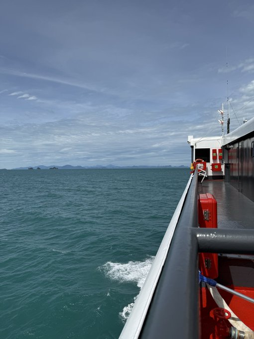 The image shows a view from a ferry boat on open water, with distant islands visible on the horizon under a partly cloudy sky, capturing the travel aspect of the author's journey. The boat features red life-saving equipment and a life ring, suggesting a sea voyage to or from an island destination—likely tied to their retreat at @islanddao.