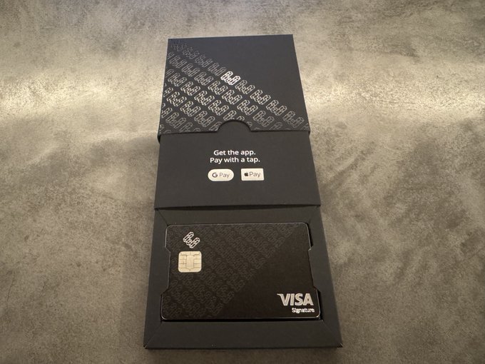 An unboxing photo of a black metal VISA Signature card from BeBank, with packaging text 'Get the app. Pay with a tap.' and Google Pay/Apple Pay logos, displayed on a gray surface.