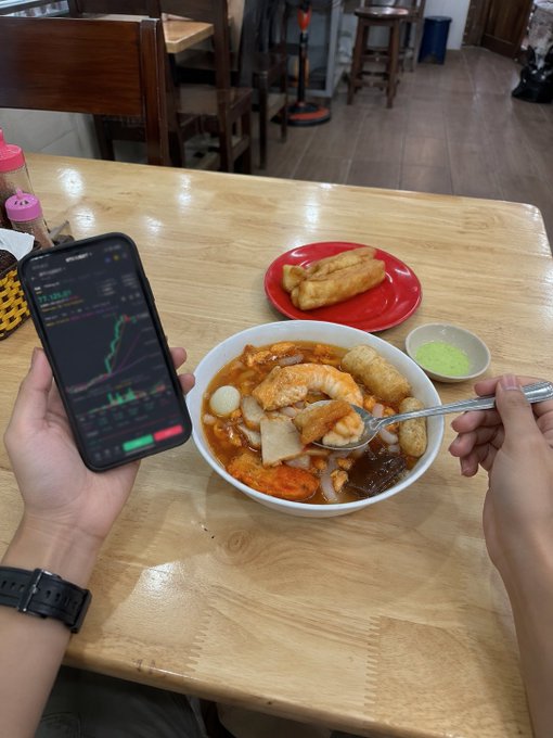 A first-person view of someone eating a bowl of soup with a spoon while checking stock charts on a smartphone, with spring rolls on a red plate nearby.