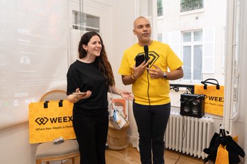 The image shows a man and woman standing next to each other, both holding cell phones. They are wearing yellow shirts with "WEEX" written on them. The background of the photo features a white wall with a window that allows natural light into the room. There is also an object in the foreground that appears to be a handbag or purse.