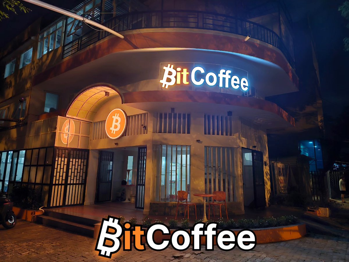 The image features an advertisement for a coffee shop called "BITCOFFEE" with a large neon sign that says "BITCOFFEE" above its entrance, which has a logo of a Bitcoin symbol. The building itself appears to be made of concrete or stone and is situated on the side of a road at night. A potted plant can also be seen in front of the coffee shop.