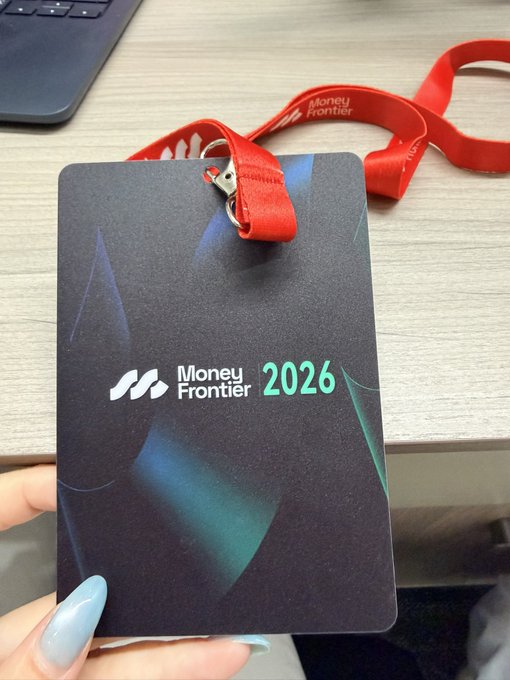 A hand with light blue nail polish holds a black "Money Frontier 2026" event badge with a red lanyard, in front of a wooden desk with a laptop. The badge represents the Money Frontier conference being promoted in Hong Kong on July 27-28, where wallet providers, stablecoin issuers, and other crypto/Web3 industry leaders are gathering.