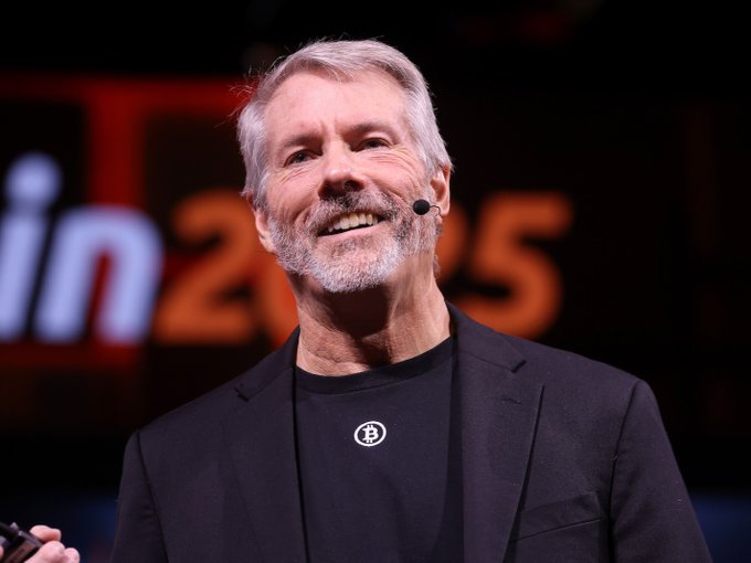 The image features an older man wearing a suit, standing on stage with a microphone in front of him. He has a beard and appears to be smiling at the camera. The background shows a large screen displaying the text "LATEST: Michael Saylor predicts Bitcoin will recover by year-end and reach a new all-time high within 12 months." This suggests that the man is giving a speech or presentation related to Bitcoin, possibly discussing its potential future value or market trends.