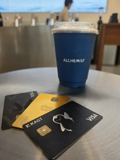 A blue "ALCHEMIST" branded cup sits on a metal table next to three bank cards, including a black "KAST" Visa card featuring a white duck logo.