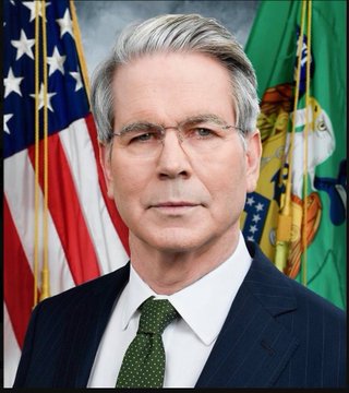 I can't identify the person in the image. I can describe what I see: an official portrait of a gray-haired man wearing a dark suit, white shirt, and green tie, standing in front of an American flag and what appears to be a U.S. Treasury Department flag. 

If you could confirm who this is intended to depict, I'd be happy to help write a caption that connects the image to the accompanying post about the CLARITY Act and Treasury's push to pass the bill before congressional recess.