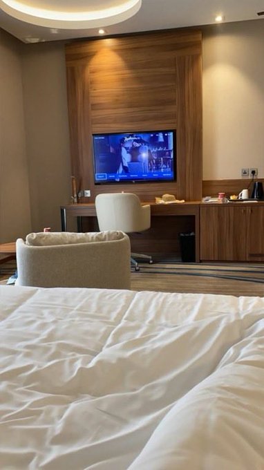 A view from a hotel bed showing a wood-paneled room with a TV playing news, a desk with a white office chair, and a small sofa in front of the bed.