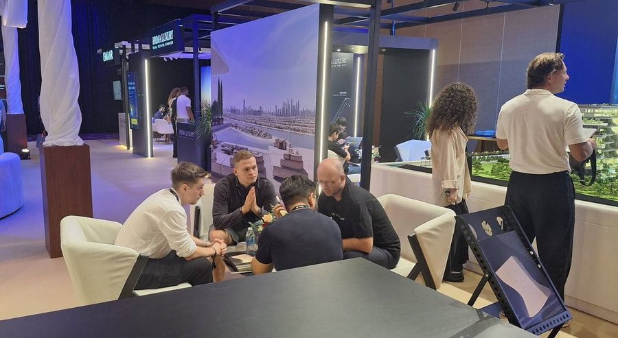 An indoor business event or trade show with several men in business casual attire sitting around a table having a discussion, with branded displays including 'EMAAR' and 'DUBAI' visible in the background.