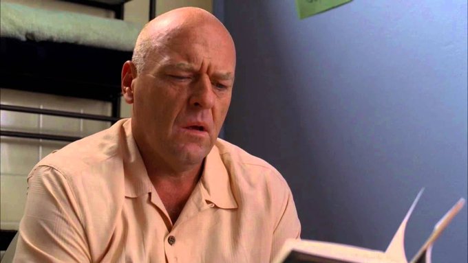 Dean Norris as Hank Schrader from Breaking Bad, wearing a beige polo shirt and grimacing while looking down.