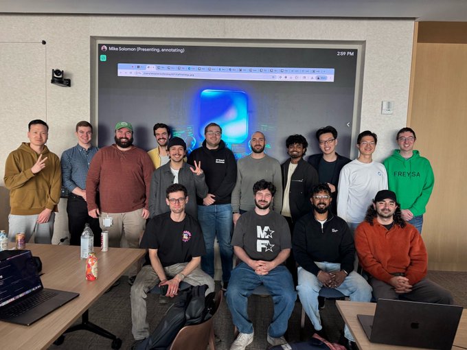 The image shows a group of people posing together for a photo, likely at an event or gathering. They are all dressed similarly, wearing black shirts and jeans. The room they are in has a large screen behind them that displays a blue background with white text on it. There is also a laptop visible in the scene.

The post caption reads: "Oh this hits hard. I'm the one in the green @freysa_ai sweater on the right about a month after joining CB. Got that sweater at the Base booth atETH Denver about a month after joining CB. I mainly went to the hackathon for the free pizza and to make friends in NYC (and to hang out with @mykcryptodev".