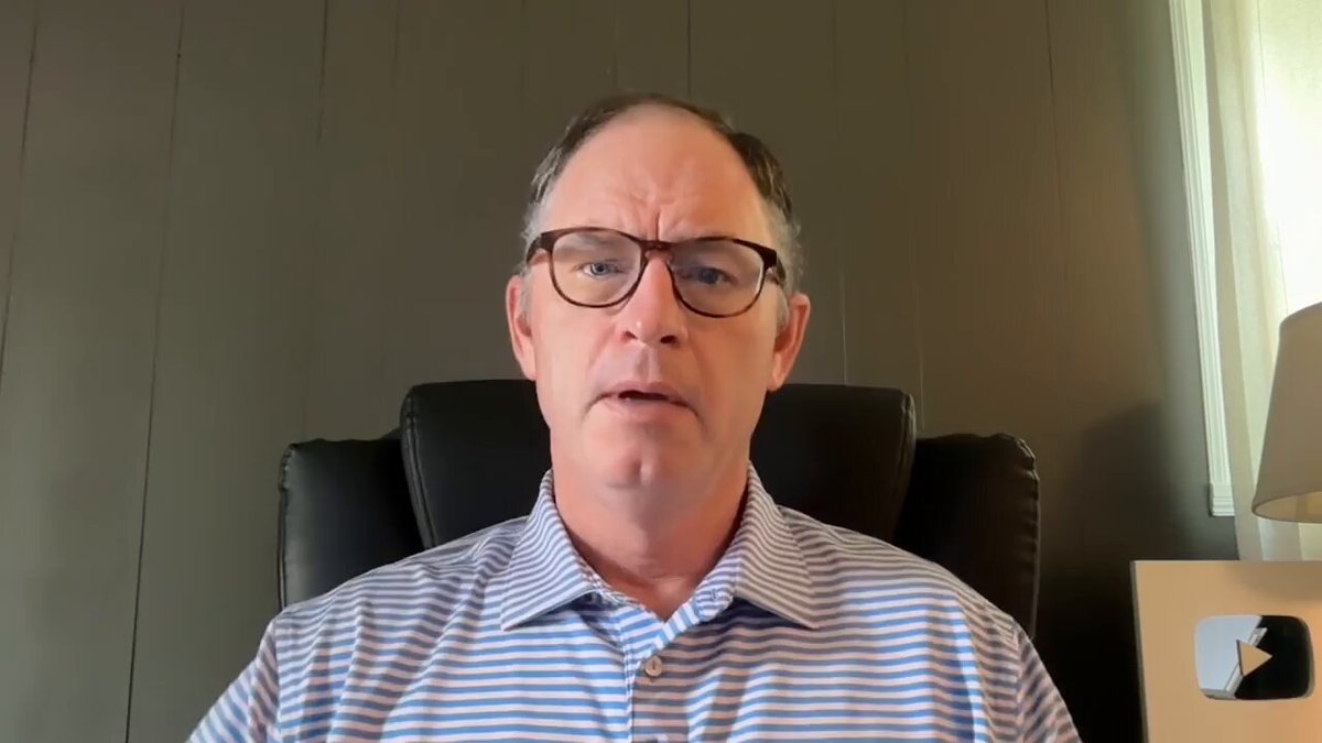 The image shows a man wearing glasses sitting in an office chair, looking at the camera with a serious expression. There are two lamps visible in the background, one near the center of the room and another towards the right side. The scene appears to be set in an office environment where the man is likely working or attending a meeting.