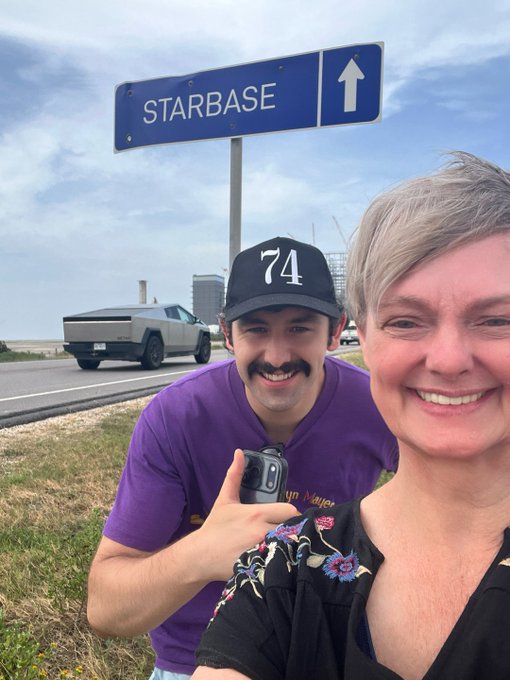 A selfie of a smiling man and woman standing in front of a blue road sign reading 'STARBASE' with an upward arrow, with a Cybertruck visible in the background.